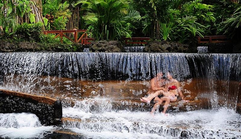 A couple relaxing under a small waterfall in empty hot springs in Costa Rica. 