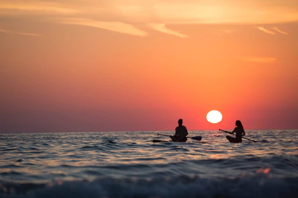 A couple sitting on paddle boards on the ocean as the sun sets behind them